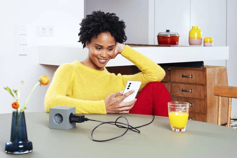 Junge Frau sitzt mit Samsung Schnellladegerät am Tisch und lächelt in ihr Smartphone.