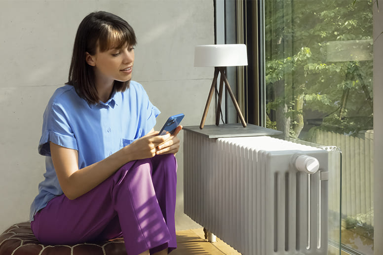 Person schaut auf ein Handy und steht vor der Heizung mit smartem Thermostat.