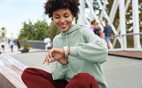 Eine junge Frau mit grünem Pulli und roter Hose sitzt draußen auf einer Mauer und blickt gut gelaunt auf ihre Smartwatch