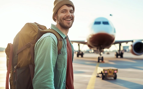 Ein Mann lächelt in die Kamera, er steht auf einem Flugfeld, im Hintergrund steht ein Flugzeug. Der Mann trägt eine Strickmütze, Rucksack, Hemd und T-shirt.