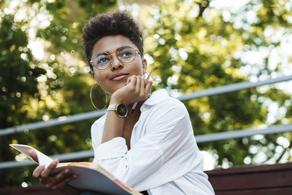 Eine Frau im Business-Look sitzt draußen auf einer Bank unter Bäumen und blickt verträumt an der Kamera vorbei. Am Handgelenk trägt sie die Hama Smartwatch "7000"