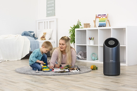 Eine junge Frau liegt zusammen mit einem Kind auf dem Boden im Kinderzimmer und spielt. Daneben steht der Hama Luftreiniger "Smart"