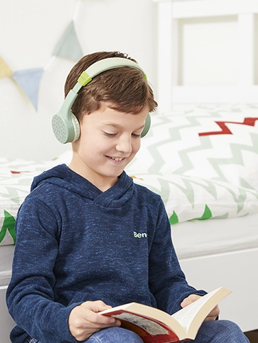 Ein Junge sitzt in seinem Zimmer und liest ein Buch, während er den Hama Bluetooth®-Kinderkopfhörer "Teens Guard" trägt