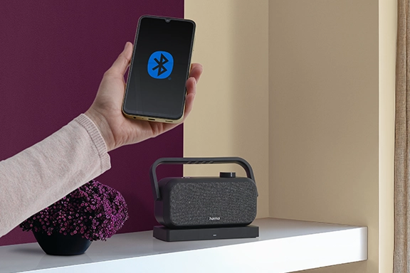 Eine Hand hält ein Smartphone mit Bluetooth-Logo auf dem Display ins Bild. Im Hintergrund steht der Lautsprecher für Fernseher auf einem weißen Sideboard.