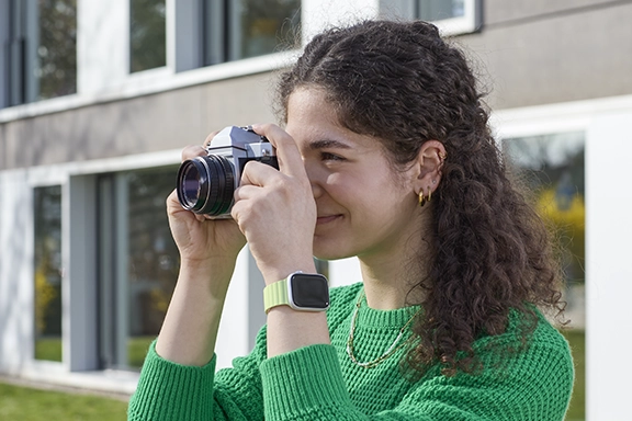 Eine junge Frau in einem grünen Strickpullover trägt eine Apple Watch zusammen mit dem Hama Magnetarmband "Fantastic Feel" und fotografiert mit einer Retrokamera ihre Umgebung.