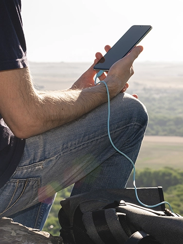 Mann sitzt beim Wandern in auf einem Hügel und lädt sein Smartphone mit dem Hama Power Pack "Supreme 10HD"